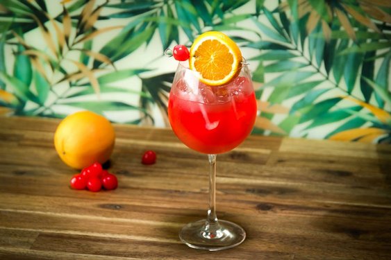 Das Bild zeigt einen roten Cocktail in einem Weinglas mit Orangendeko